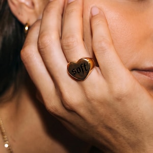 May include: A gold-toned heart-shaped ring featuring the word soft engraved in a black gothic-style font. The ring has a smooth, polished surface and is worn on the ring finger, showcasing a simple band design that encircles the finger.