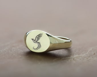 May include: A gold signet ring with an oval-shaped top featuring a black and white engraved design of a bird in flight.
