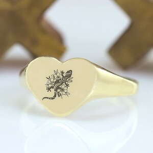 May include: A gold heart-shaped signet ring with a black and white engraved design of a gecko with flowers.