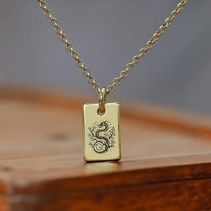Puede incluir: Collar de cadena dorada con un colgante rectangular con una imagen grabada de un dragón.