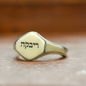 Puede incluir: Un anillo de sello de oro con una cara hexagonal grabada con la palabra hebrea "רבקה" (Rebekah).