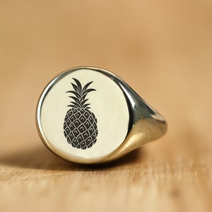 Könnte beinhalten: Ein silberner Siegelring mit einem eingravierten schwarzen Ananas-Motiv auf der Vorderseite.