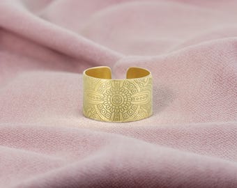 Könnte beinhalten: Ein goldfarbener, verstellbarer Ring mit einem eingravierten Blumenmuster. Der Ring hat ein breites Band mit offenem Design. Der Ring wird auf einem rosa Stoffhintergrund präsentiert.