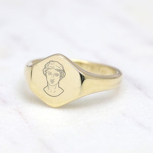 May include: A gold signet ring with a raised, octagonal face featuring a line drawing of a woman's head.