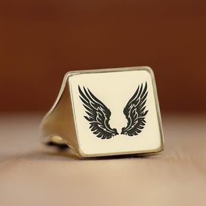 May include: A silver ring with a square white face featuring a black silhouette of angel wings.