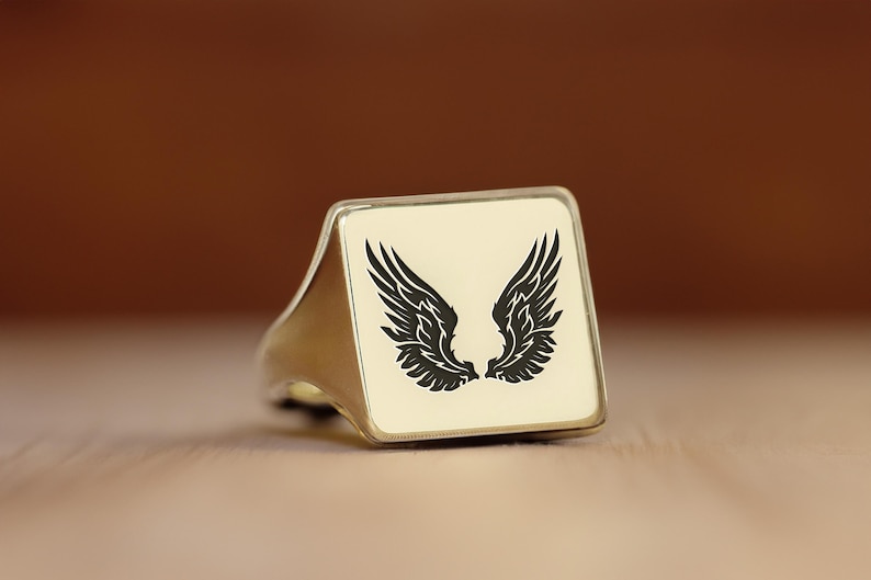May include: A silver ring with a square white face featuring a black silhouette of angel wings.