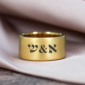 May include: A wide, gold-colored ring with engraved Hebrew letters. The ring has a brushed finish and sits on a soft, pinkish-beige fabric. The letters are dark and stand out against the gold.