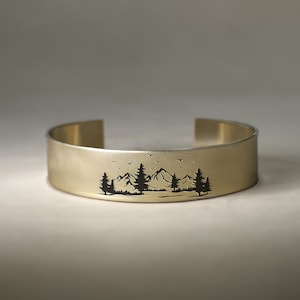 May include: A gold-toned metal bracelet with a minimalist design featuring a silhouette of a mountain range and trees.
