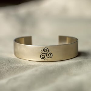 May include: A gold metal cuff bracelet with a Celtic knot design engraved on the front.