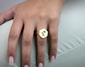 May include: A gold signet ring with a hexagonal face. The ring features a black palm tree design. The ring is worn on a finger. The background is a light green colour.