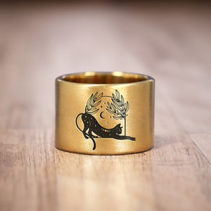 May include: A gold ring featuring a black cat design. The cat is lying down with its head turned to the side. The design also includes a crescent moon and leaves.