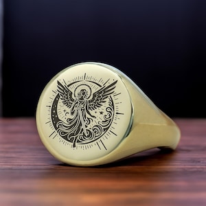 May include: A gold signet ring with a black and white engraved design of an angel with wings, surrounded by a crescent moon and stars.
