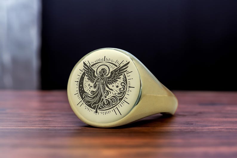 May include: A gold signet ring with a black and white engraved design of an angel with wings, surrounded by a crescent moon and stars.