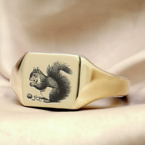 May include: A gold signet ring with a square face featuring an engraved image of a squirrel holding a nut.