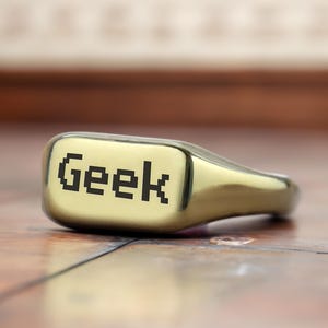 May include: A gold-colored ring with the word "Geek" inscribed in black pixelated letters.
