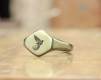 May include: A silver signet ring with a square face featuring a black engraved design of a stylized bird.