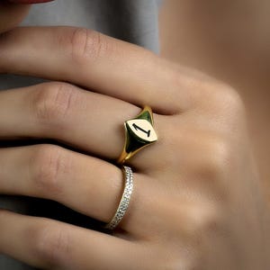 May include: A gold signet ring with the number "1" in black on a white background. A smaller gold band with small, clear stones is also visible. The rings are on a hand with pink nail polish.