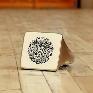 May include: A silver ring with a square face featuring a black and white image of a cobra with a mandala design.