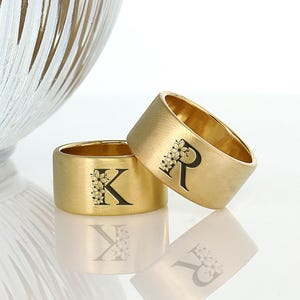 Könnte beinhalten: Zwei goldene Eheringe mit gebürsteter Oberfläche. Die Ringe sind mit den Buchstaben "K" und "R" graviert, die von kleinen floralen Designs umgeben sind.