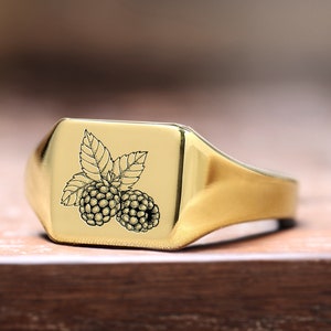 May include: A gold signet ring with a square face featuring an engraved image of two blackberries and leaves.