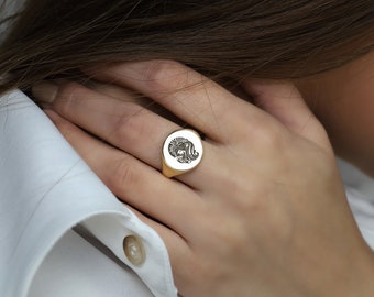 May include: A gold signet ring featuring a detailed engraved design of a woman's face. The ring is oval shaped and is worn on a person's finger.