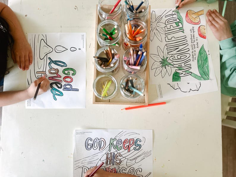 May include: Two children color on a white table with colorful pencils. The table has a set of jars filled with colored pencils and three coloring pages. The coloring pages have the phrases "God loves", "Magnified", and "God keeps his promises".