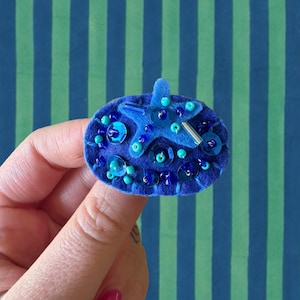 May include: An oval-shaped blue felt brooch featuring a central blue felt starfish appliqué. The surface is decorated with a mix of dark blue and turquoise sequins, small round beads, and a single silver-toned cylindrical bead accent.