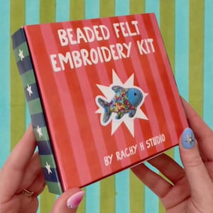 May include: A square box for a beaded felt embroidery kit featuring red and pink vertical stripes. The text reads BEADED FELT EMBROIDERY KIT BY RACHY H STUDIO. A blue fish illustration with colorful beads is centered on a white starburst shape.