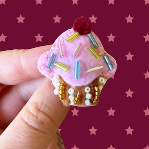 Può includere: Una piccola spilla a forma di cupcake realizzata in feltro rosa con un pompon rosso scuro sulla parte superiore. La glassa è decorata con varie perline di vetro tubolari colorate. La base è in feltro marrone decorata con perline bianche e oro.