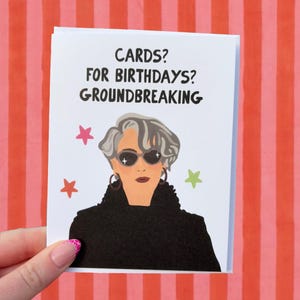 Pode incluir: Um cartão branco com o texto "CARDS? FOR BIRTHDAYS? GROUNDBREAKING" e uma ilustração de uma mulher usando óculos de sol e um casaco preto. O cartão é segurado contra um fundo listrado vermelho e branco.