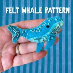 May include: A bright blue felt whale ornament featuring the text FELT WHALE PATTERN. The whale is decorated with clear and silver beads, sequins, and embroidery stitches, including a small black bead for the eye and a fin on the underside.