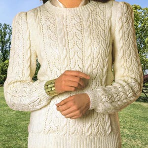 May include: Cream-colored knitted sweater with intricate cable and leaf patterns. The long-sleeved sweater features a crew neck and ribbed cuffs and hem. The sleeves have a slight puff at the shoulders. A green and gold bracelet is visible on the left wrist.