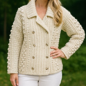May include: Cream-colored, double-breasted knit jacket with a notched collar and gold-tone buttons. The jacket features a textured stitch pattern and is worn with white pants. A woman is wearing the jacket and smiling.