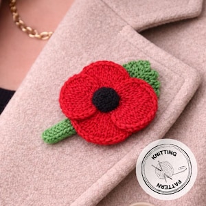 Può includere: Una spilla a forma di fiore di papavero rosso lavorato a maglia, con centro nero e foglie verdi, è attaccata a un cappotto beige. La spilla è fatta a mano e ha un aspetto strutturato. È visibile un adesivo rotondo con la scritta "Knitting Pattern".