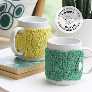 Könnte beinhalten: Zwei weiße Keramikbecher mit gehäkelten Bezügen. Ein Bezug ist gelb, der andere grün. Der grüne Bezug hat einen Holzknopf. Das Bild enthält ein "CROCHET PATTERN"-Logo. Die Becher stehen auf einer weißen Oberfläche.