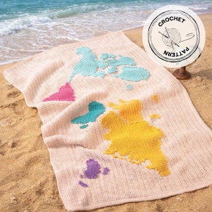 Può includere: Una coperta all'uncinetto con un design a mappa del mondo, che mostra i continenti in rosa, blu, giallo e viola. La coperta è appoggiata su una spiaggia sabbiosa con il mare sullo sfondo. È visibile anche un timbro circolare con "CROCHET PATTERN".