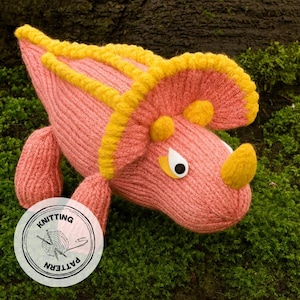 May include: A knitted pink Triceratops dinosaur with yellow accents on its frill and horn. The dinosaur has white and black eyes and is set against a mossy background. A circular "Knitting Pattern" logo is in the lower left.