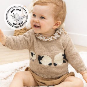 Op de afbeelding: Een lachende baby draagt een beige gebreide trui met een schapenontwerp en een bloemenkraag. De trui heeft lange mouwen en wordt gecombineerd met bruine shorts. Een cirkelvormige afbeelding met de woorden "Knitting Pattern" en een bol wol staat op de achtergrond.