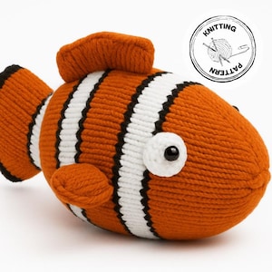 May include: A knitted orange and white clownfish toy with black stripes and a white eye. The fish has a small fin on top and a tail fin. The image includes a circular logo that says "Knitting Pattern".