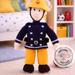 Może przedstawiać: Dzianinowa lalka przebrana za strażaka, w żółtym kasku, granatowej kurtce z białymi kropkami i żółtych spodniach. Lalka ma czarne buty i czarny pasek. Okrągła naklejka z napisem "Knitting Pattern" znajduje się na pierwszym planie.