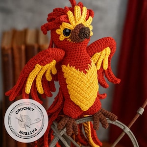 Puede incluir: Un fénix tejido a mano en crochet, en rojo vibrante, amarillo y marrón. El ave presenta alas detalladas, un pecho amarillo y una cresta. La imagen incluye un logotipo de "CROCHET PATTERN", lo que sugiere un proyecto de manualidades o un artículo hecho a mano.