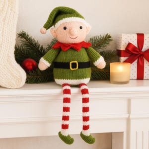 Puede incluir: Muñeca de elfo navideño tejida, con un atuendo verde, cuello rojo y piernas a rayas rojas y blancas. El elfo tiene un gorro verde, un cinturón negro con hebilla dorada y una cara sonriente. La muñeca está sobre una superficie blanca.