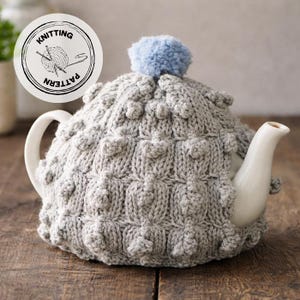 Puede incluir: Una tetera de cerámica blanca cubierta con un cubrete de té de punto gris. El cubrete tiene un patrón texturizado y un pompón azul claro en la parte superior. Una etiqueta circular con las palabras "Knitting Pattern" es visible.