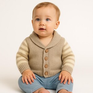 May include: A baby wearing a tan knit cardigan with a shawl collar and wooden buttons. The sleeves have horizontal stripes. The baby is also wearing light blue pants.