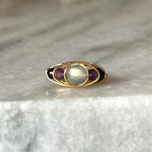 May include: A gold-toned ring featuring a central, round, iridescent stone, flanked by two purple stones and two black stones. The stones are set within a woven gold wire design. The ring is displayed on a light-colored surface.