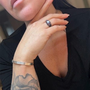 May include: A silver cuff bracelet with the inscription "live your box". A black ring with a light blue stone and a dark metal band is on a finger. A black v-neck shirt is worn.