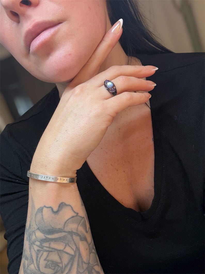 May include: A silver cuff bracelet with the inscription "live your box". A black ring with a light blue stone and a dark metal band is on a finger. A black v-neck shirt is worn.