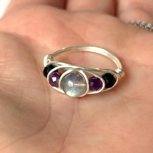 May include: A handmade silver wire ring featuring a central iridescent stone, flanked by two purple and two black beads. The ring's design incorporates a woven wire pattern, creating a unique and artistic piece of jewelry.