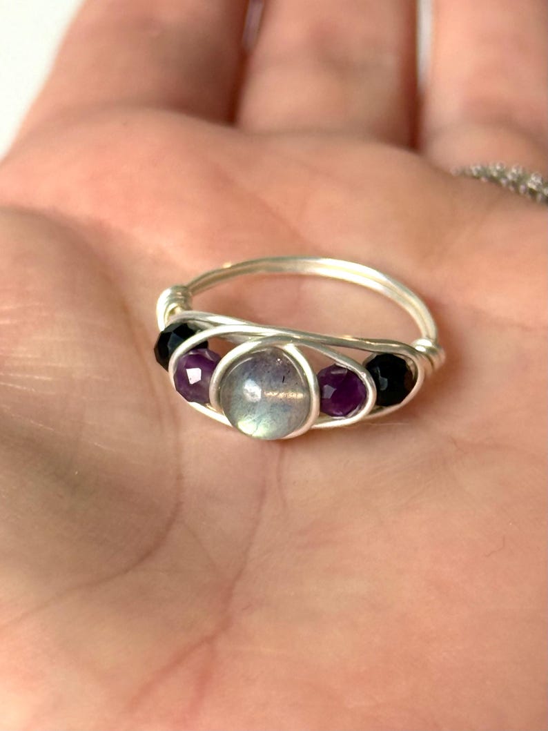 May include: A handmade silver wire ring featuring a central iridescent stone, flanked by two purple and two black beads. The ring's design incorporates a woven wire pattern, creating a unique and artistic piece of jewelry.