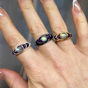 May include: Three wire-wrapped rings with gemstone accents. The rings feature a central, iridescent stone flanked by smaller purple stones. One ring has a silver wire, another black, and the third is gold. The rings are displayed on a hand.